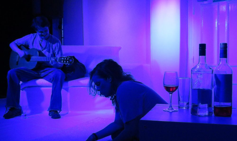 A photo of a performance at a pub theatre showing two people on stage, one sat on a sofa playing guitar the other sitting on the floor next to a glass of wine, with purple lighting